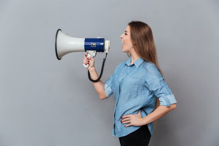 Mulher de camisa azul falando energicamente em um megafone, representando a comunicação direta.