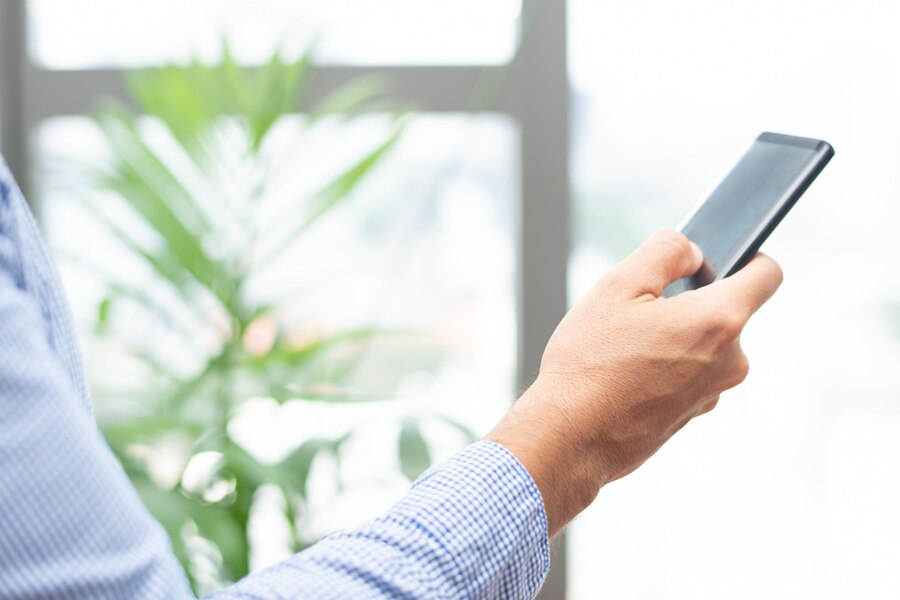 Homem segurando smartphone com uma mão, fazendo um gesto de clique, em um ambiente de escritório com uma planta ao fundo.