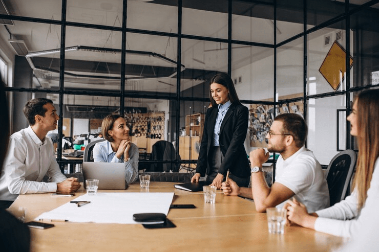 Equipe corporativa diversificada participando de uma reunião de estratégia em um ambiente de trabalho moderno e arejado. Uma mulher de pé lidera a discussão com colegas focados ao redor de uma mesa com laptops, simbolizando colaboração, liderança feminina e engajamento profissional em espaços de coworking.