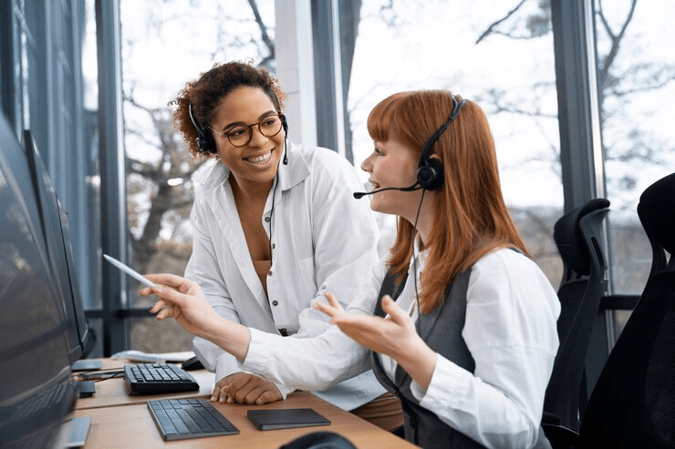 Duas pessoas sentadas em uma mesa, usando fones de ouvido e olhando para a tela de um computador. Parece ser um ambiente de trabalho colaborativo, possivelmente em um ambiente de atendimento ao cliente ou suporte técnico.