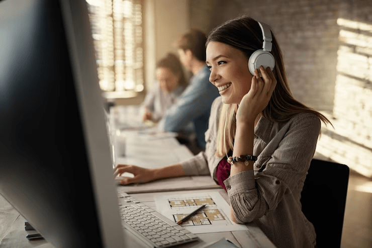 Uma pessoa com fones de ouvido sentada em frente a um computador, aparentemente envolvida em uma tarefa, possivelmente ouvindo algo ou em uma reunião virtual. A configuração sugere foco em tarefas individuais, possivelmente em uma casa ou escritório com planta aberta.