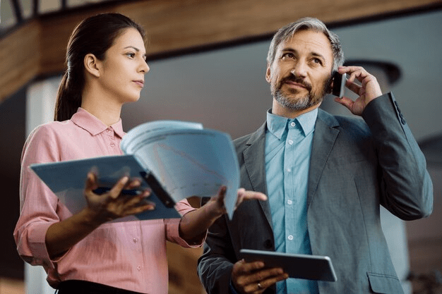 Dois profissionais em ambiente empresarial. Um deles é um homem que parece estar conversando ao celular, vestido de terno, olhando para cima, possivelmente ouvindo ou pensando. A outra é uma mulher segurando uma pasta ou fichário aberta, aparentemente interagindo com o homem, talvez fornecendo informações para a discussão.