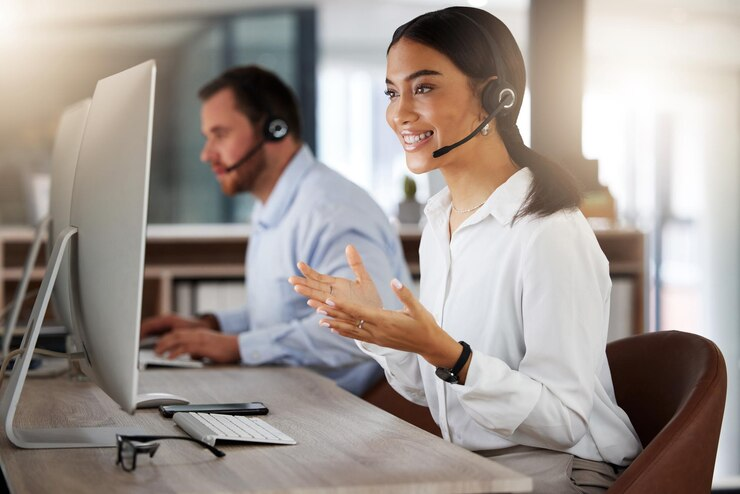 Operadora de telemarketing com fone de ouvido e microfone sorrindo, gesticulando com as mãos, com colega ao fundo, exemplificando um eficaz pós atendimento.