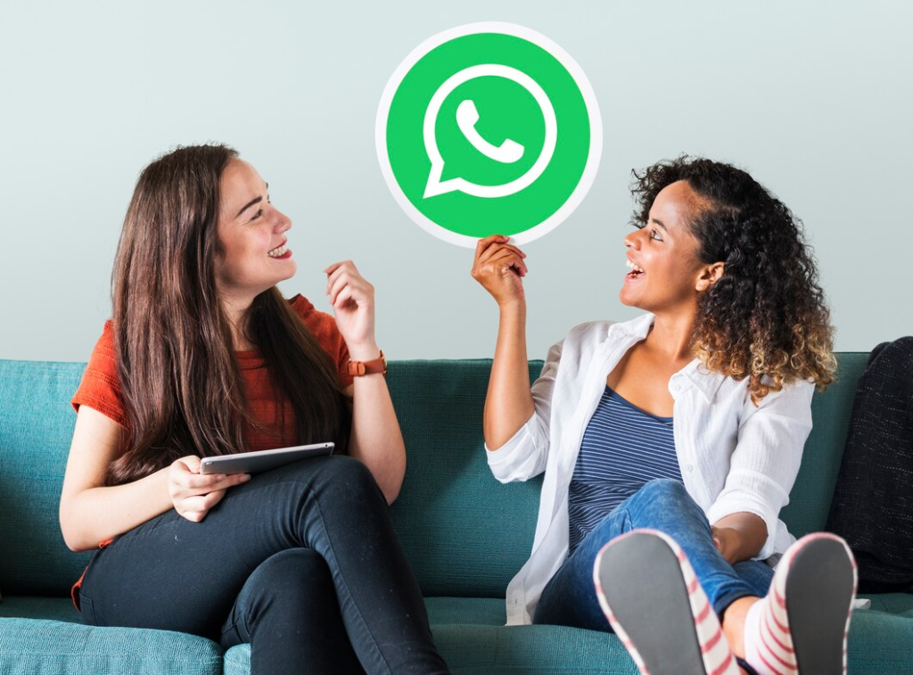 Duas amigas estão tendo uma conversa envolvente em um sofá com um ícone do WhatsApp flutuando acima. A imagem sugere a facilidade e simpatia de usar o WhatsApp para comunicação