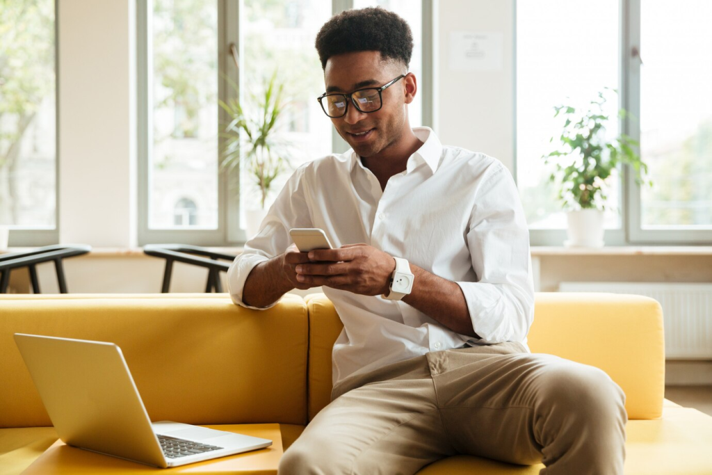 Homem de negócios utilizando o WhatsApp em seu smartphone enquanto trabalha em um laptop, evidenciando a multifuncionalidade do aplicativo no ambiente de trabalho moderno.

