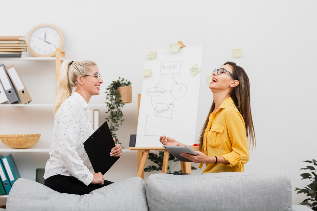 Duas colegas de trabalho em um espaço de escritório leve e arejado, sorrindo e engajadas em uma discussão produtiva em frente a um flip chart com notas adesivas e gráficos. Elas representam o espírito de parceria e inovação no local de trabalho.