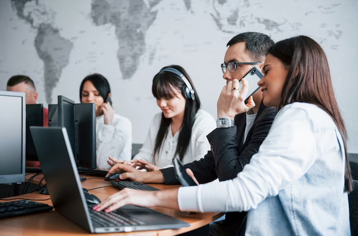 Um grupo de profissionais de atendimento ao cliente trabalhando com computadores e headsets em um ambiente de escritório, com um mapa-múndi ao fundo, representando a natureza global do suporte ao cliente moderno.





