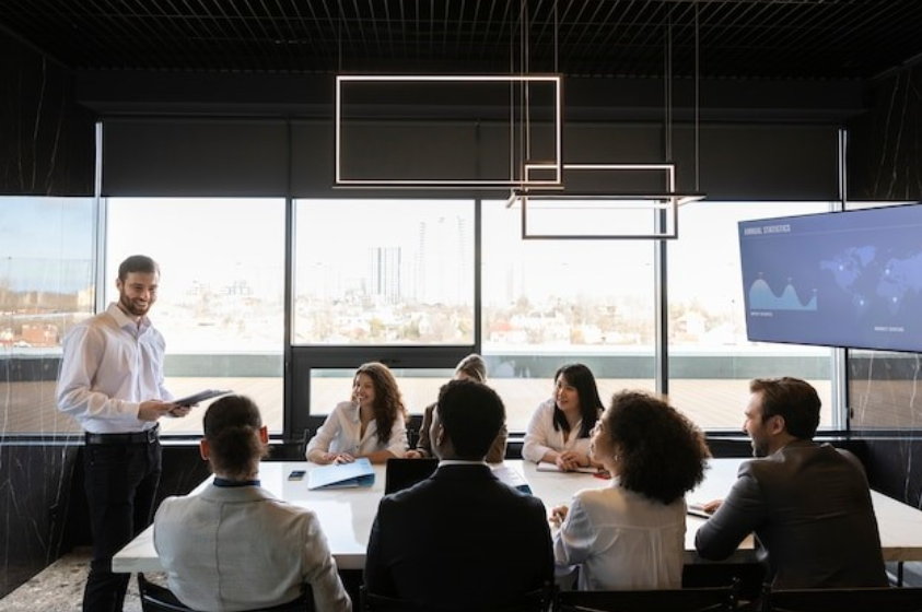 Profissional conduzindo uma apresentação em uma sala de reuniões moderna com colegas atentos e gráficos exibidos em um monitor.