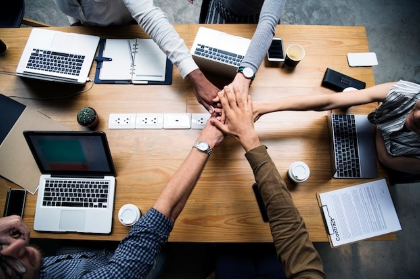 Equipe de trabalho colocando as mãos juntas em gesto de colaboração sobre uma mesa com laptops e documentos.