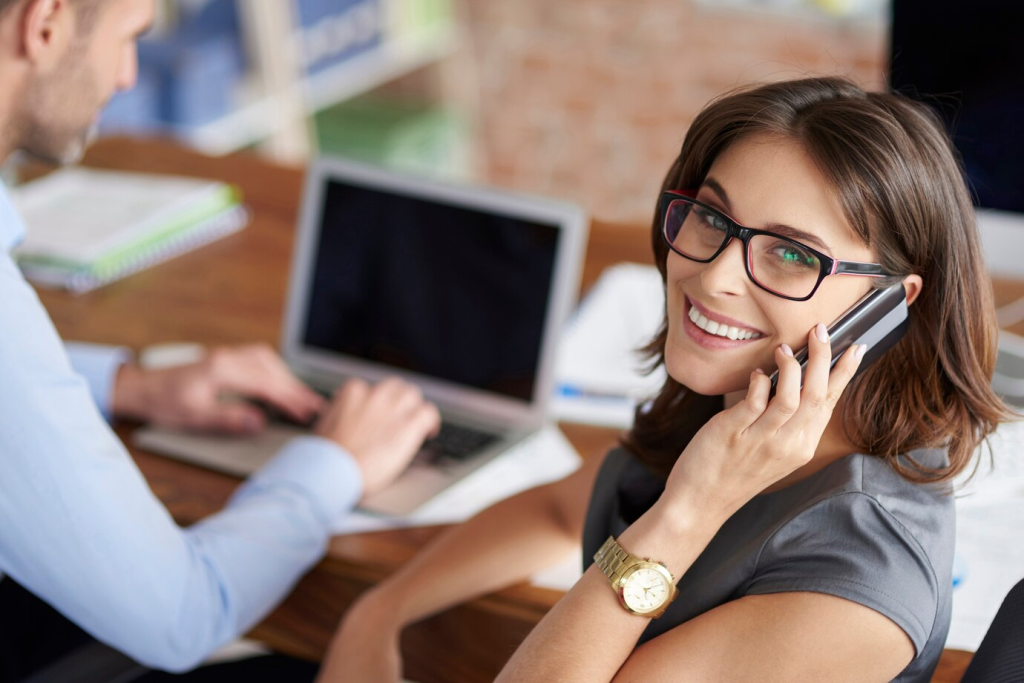 Mulher de negócios sorridente falando ao telefone enquanto trabalha ao lado de um colega masculino que digita em um laptop em um ambiente de escritório moderno.