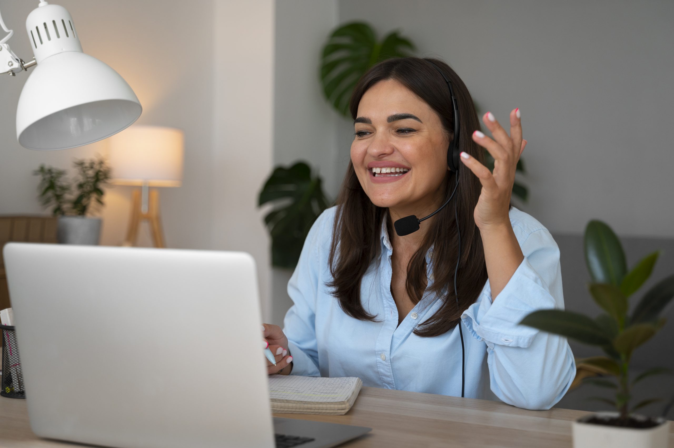 A imagem mostra uma mulher sorridente, usando um headset, participando de uma videochamada em casa. Ela está sentada à mesa com um laptop à sua frente, um bloco de notas e uma caneta na mão. O ambiente ao redor é acolhedor, com plantas e iluminação suave, criando um espaço de trabalho confortável e produtivo.